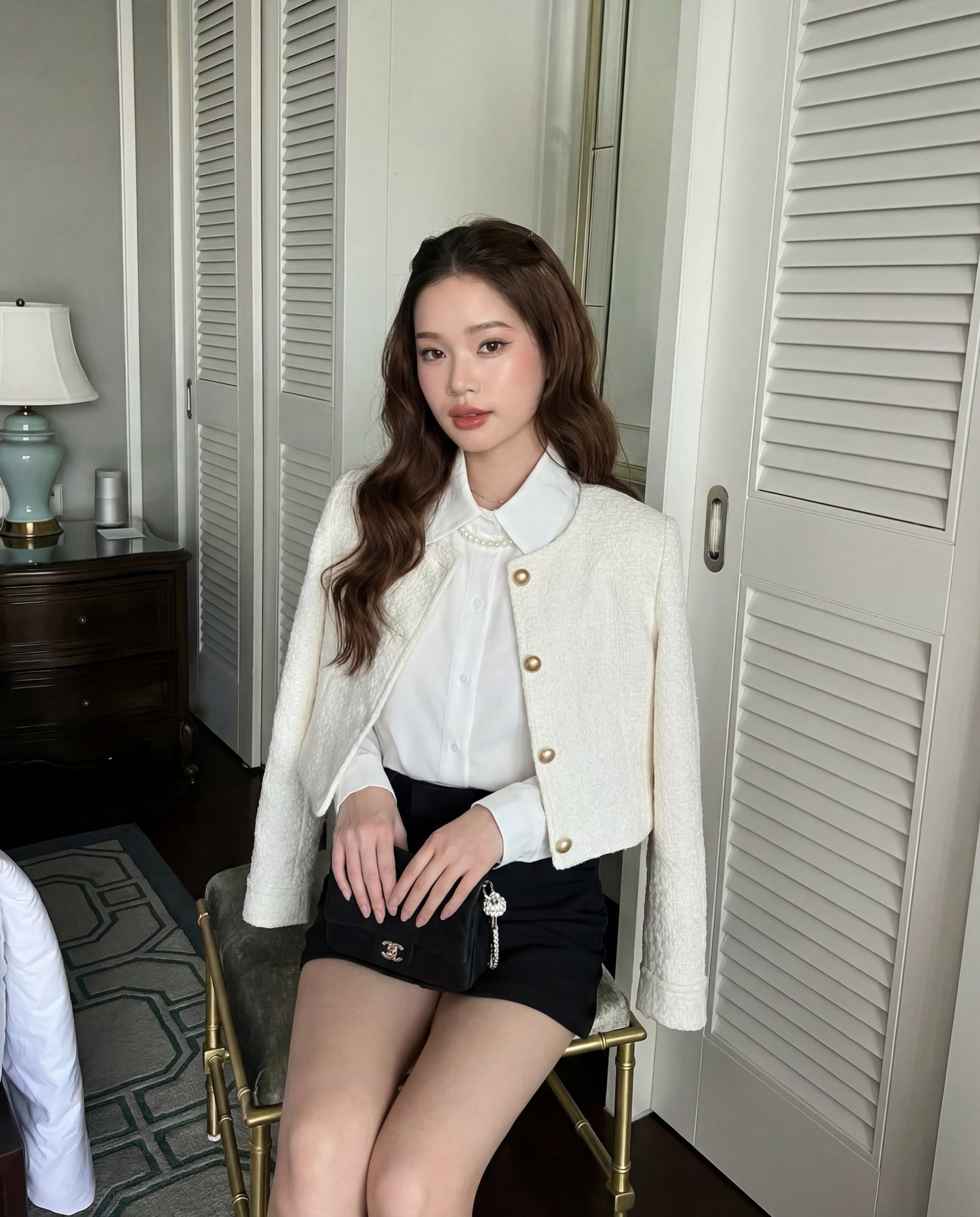 一位棕色長髮的女士坐在椅子上，身穿.lookbook.select Puff Sleeve Shirt 🖤新品優惠2件9折、乳白色外套、黑色短裙，手上拿著一個小皮包，在一個有白色百葉門和床頭櫃的房間裡。.