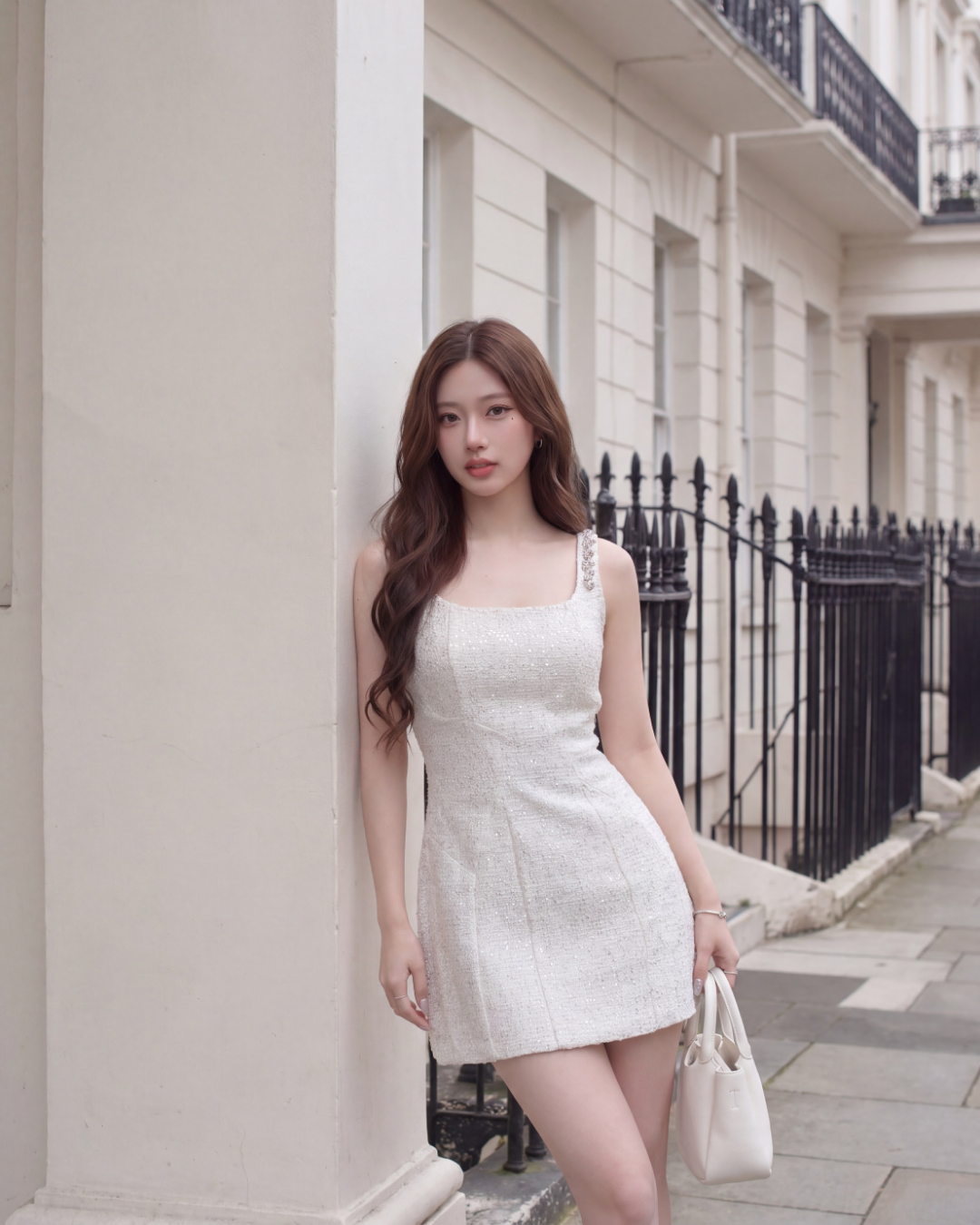 一位身穿 the.lookbook.select's Jewel Tweed Dress 🌙新品優惠2件9折的女士站在一幢輕型建築旁，手持白色手提包，背景是鐵欄杆和鋪好的人行道。.