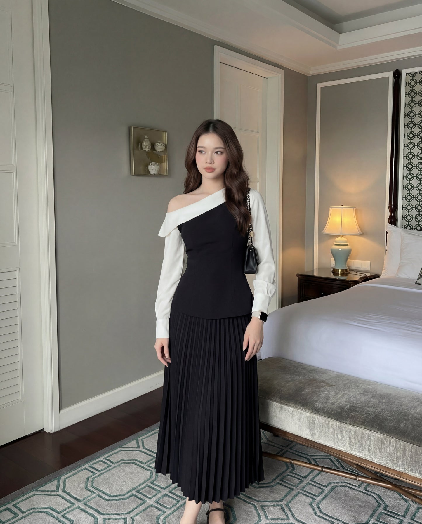 一位女士站在室內的床邊，穿著 the.lookbook.select 的 Mono Skirt Set - 百褶裙，手拿黑色手提包。.