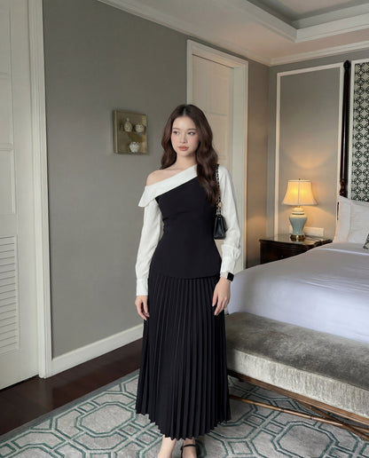 一位女士站在室內的床邊，穿著 the.lookbook.select 的 Mono Skirt Set - 百褶裙，手拿黑色手提包。.