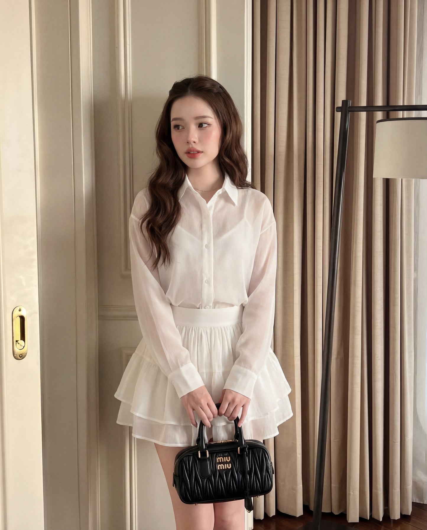 The.lookbook.select的晨霧荷葉邊套裝中的女性站在室內，手持黑色Miu Miu手袋。背景可見米色窗簾和落地燈。.