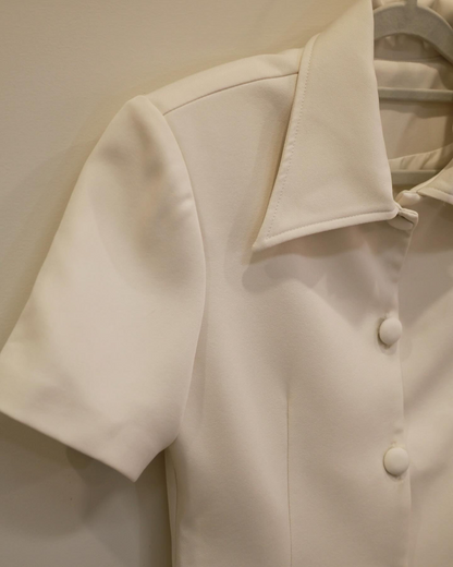 The.lookbook.select的Light Skirt Set🖤新品優惠2件9折特寫，以尖領乳白色短袖襯衫和布料包覆的鈕扣為特色，展示在米色牆壁旁的衣架上。.