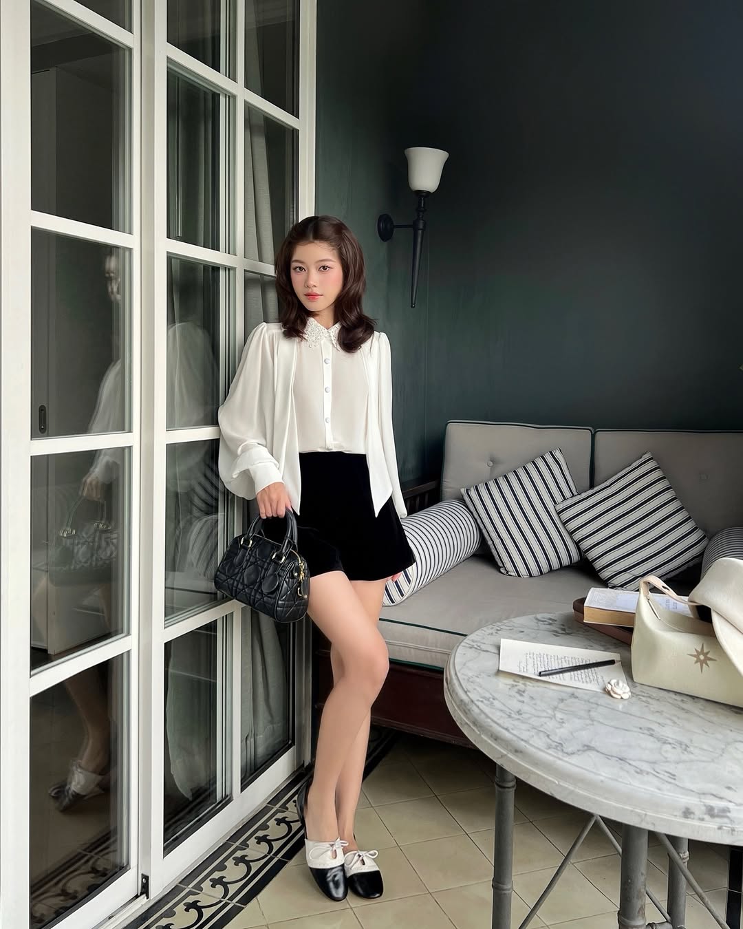 一位女士站在室內的玻璃門旁，身穿 the.lookbook.select's Heart White Shirt ❄️ 產品優惠、黑色短裙和黑色皮鞋，手拿黑色手提包；條紋沙發抱枕和大理石桌子增添了微妙的優雅氣質。.