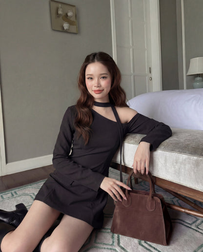 一位身穿 the.lookbook.select 單肩西裝連衣裙的女士坐在地板上，靠著長凳，手持棕色手提包，置身於現代、中性色調的房間中。.