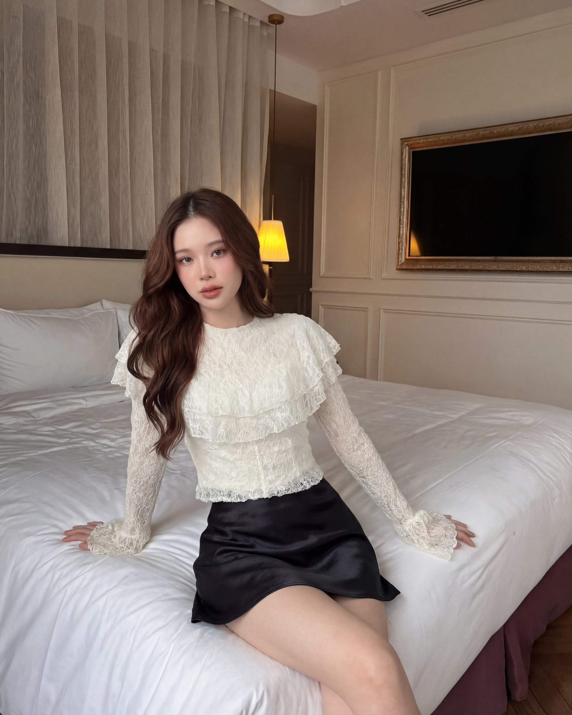 一位留著長棕髮的女子坐在一間色調中性的臥室裡的床上，身穿來自 the.lookbook.select 的「Spring Muse 蕾絲上衣」與黑色裙子；牆上可見一台電視和一幅裝裱好的照片。.