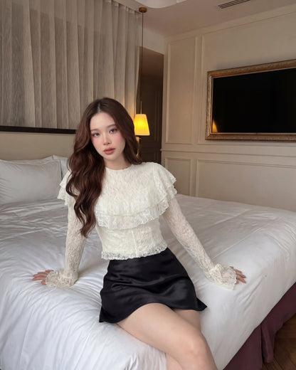 一位留著長棕髮的女子坐在一間色調中性的臥室裡的床上，身穿來自 the.lookbook.select 的「Spring Muse 蕾絲上衣」與黑色裙子；牆上可見一台電視和一幅裝裱好的照片。.