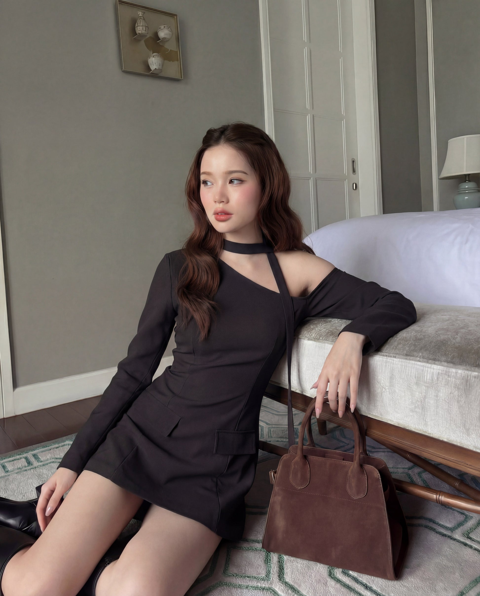 一位身穿 the.lookbook.select 單肩西裝連衣裙的女士坐在床邊的地板上，手臂靠在床上，身旁放著一個棕色手提包。.