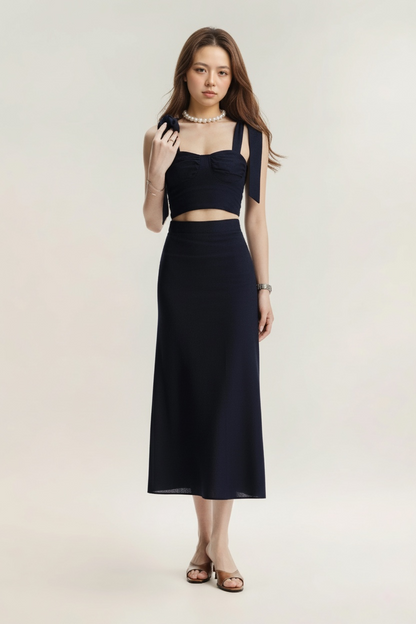 一位女士身穿 the.lookbook.select 的 Bow Ruffle Top，搭配海軍藍色長裙、珍珠項鍊、手鐲和涼鞋，以素色為背景，面朝前方。.