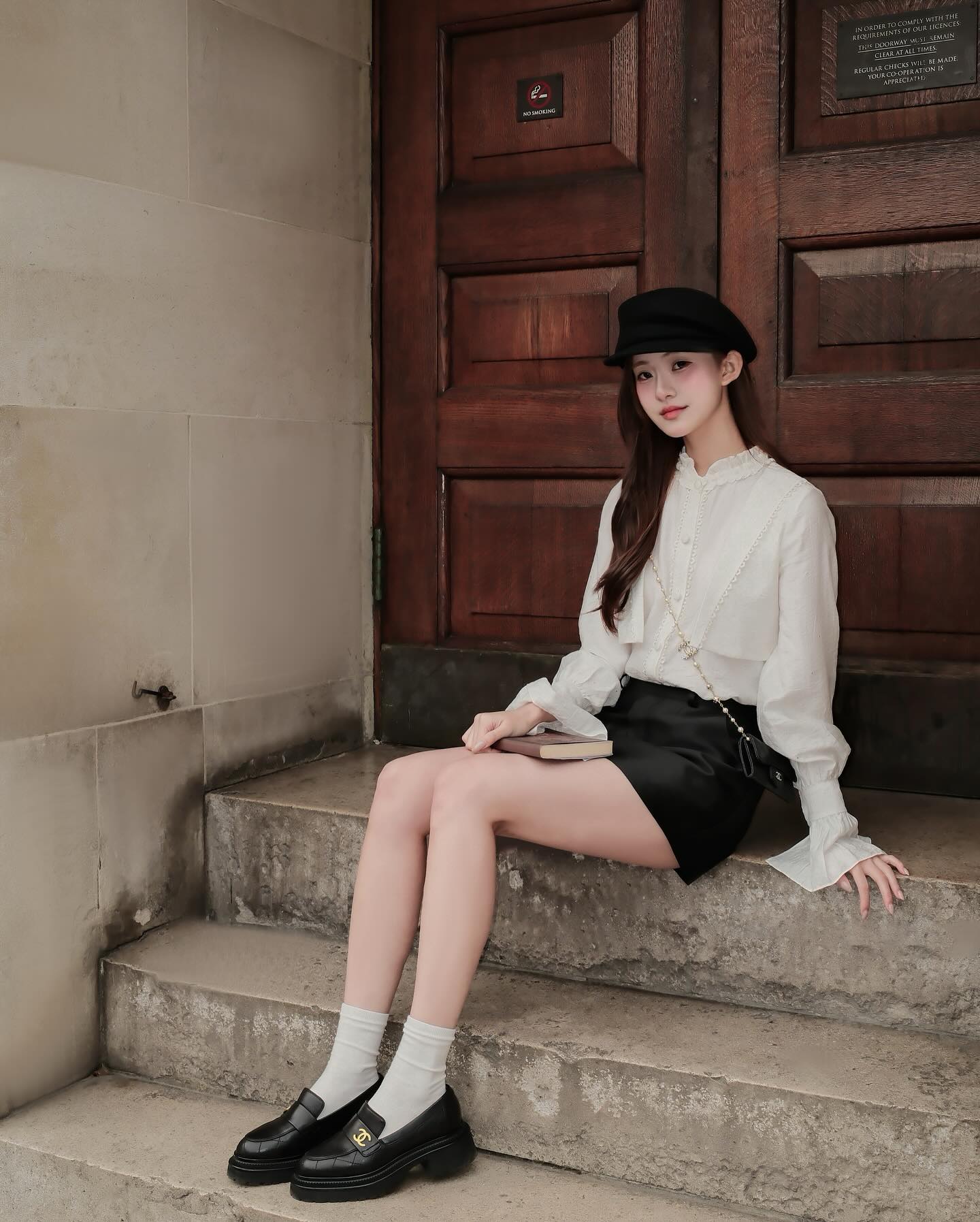一位女士穿著 the.lookbook.select 的 Hazel White Shirt，搭配黑色短裙和帽子，優雅地坐在木門前的石階上，對著鏡頭擺出法式氣質的造型。🖤新品優惠至12月5日.