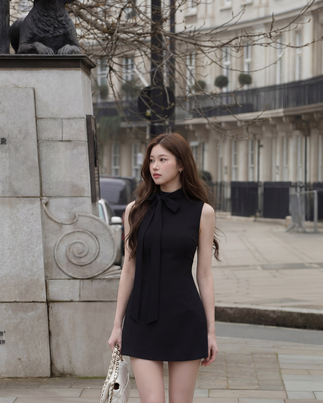 一位身穿 the.lookbook.select 的 Estate Tie-Neck Minidress 的女士站在城市街道的石碑旁，手拿白色手提包。.