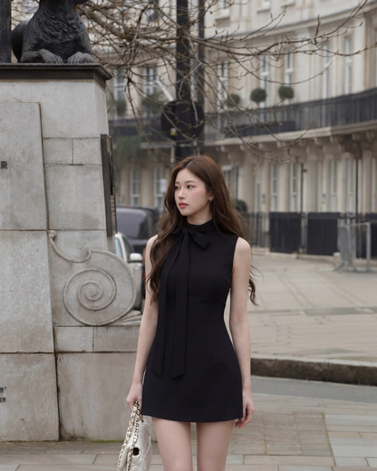 一位身穿 the.lookbook.select 的 Estate Tie-Neck Minidress 的女士站在城市街道的石碑旁，手拿白色手提包。.