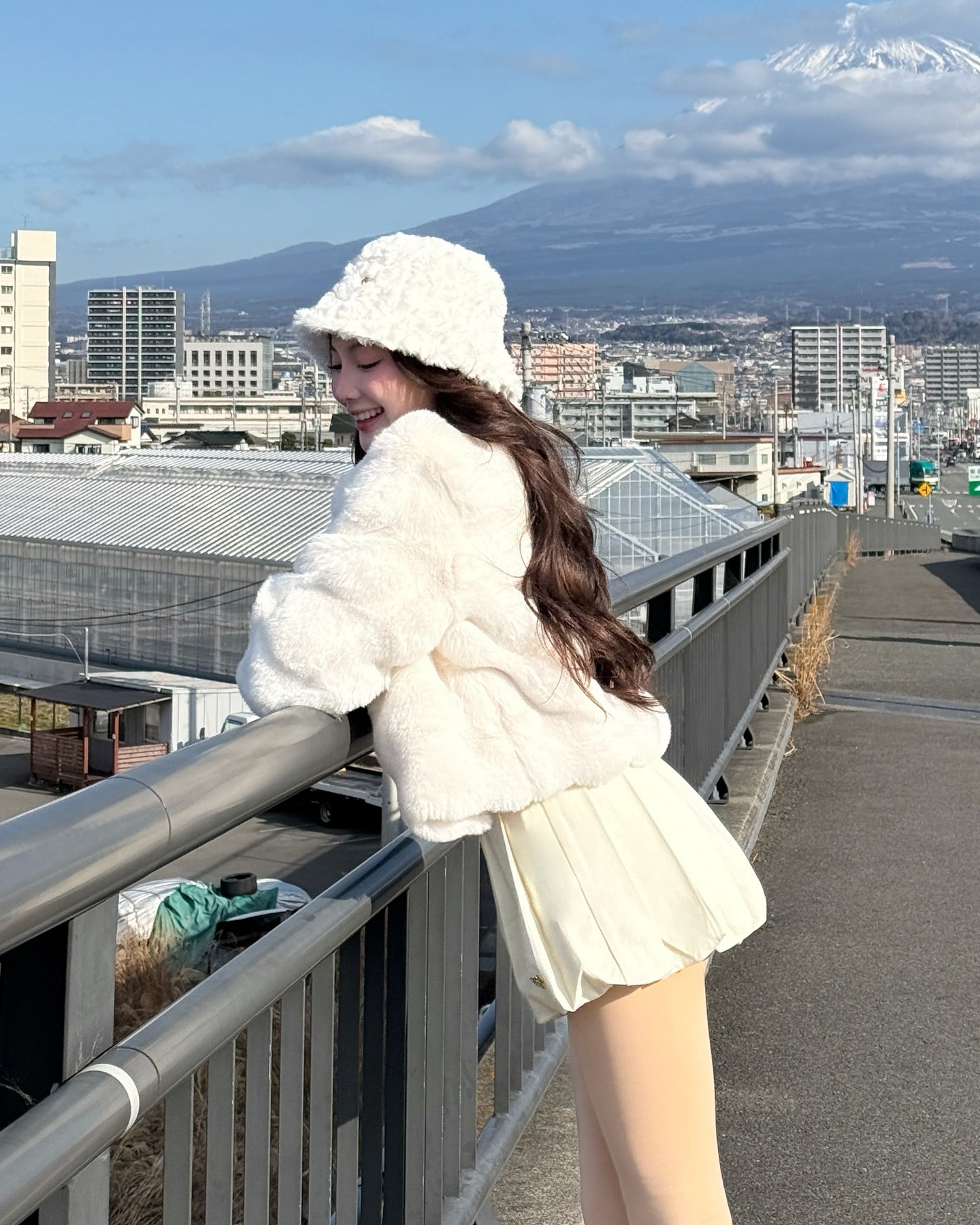 一位身穿 the.lookbook.select 的棉花糖皮草大衣 (配帽子) 的女士倚靠在欄杆上，面帶微笑，背景是城市建築和一座山。.