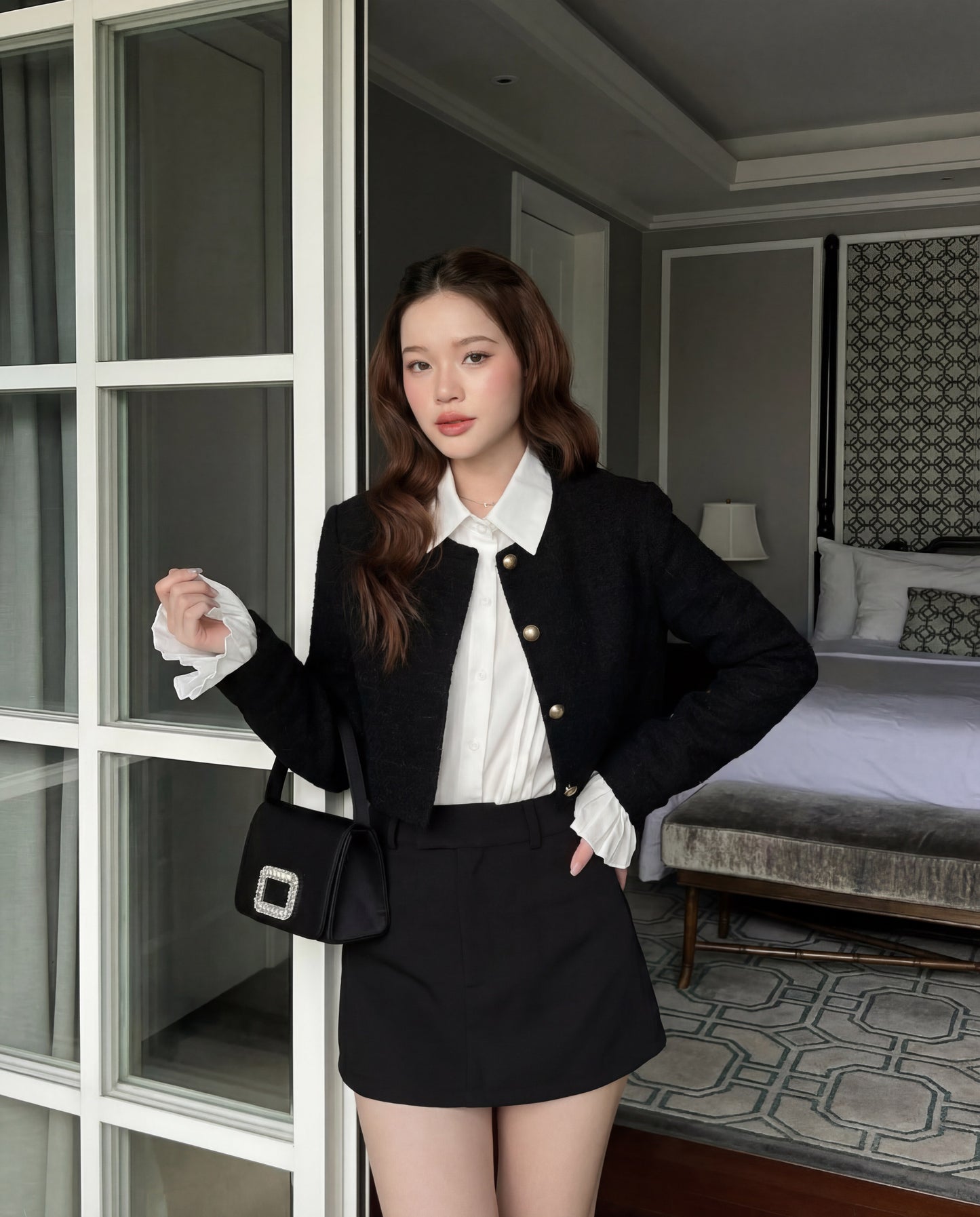一位女士站在室內，身穿 the.lookbook.select 的 Sine Tweed Coat，外搭白色襯衫和黑色短裙，手持黑色皮包。背景可見一張床和床頭櫃。.