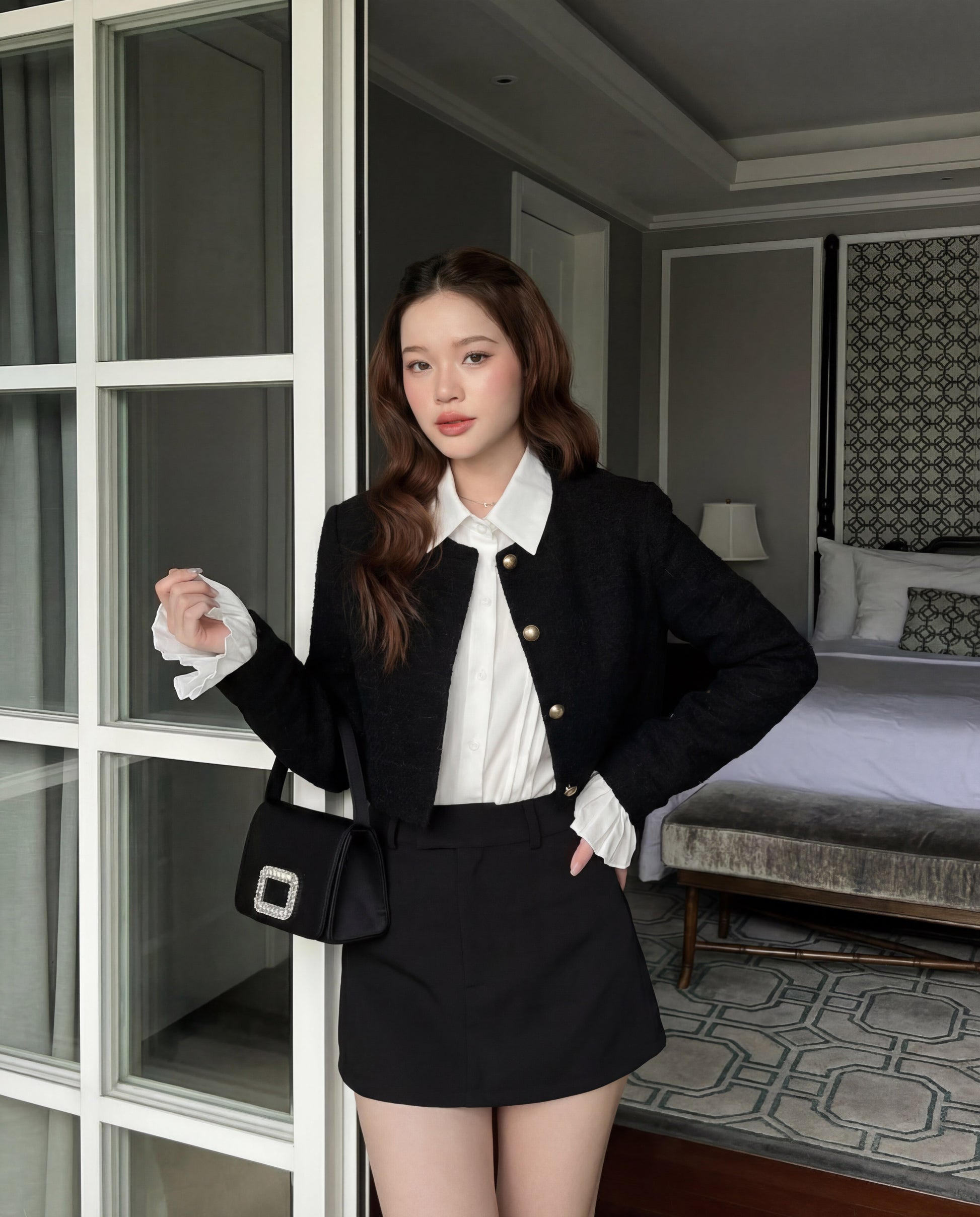 一位女士站在室內，身穿 the.lookbook.select 的 Sine Tweed Coat，外搭白色襯衫和黑色短裙，手持黑色皮包。背景可見一張床和床頭櫃。.