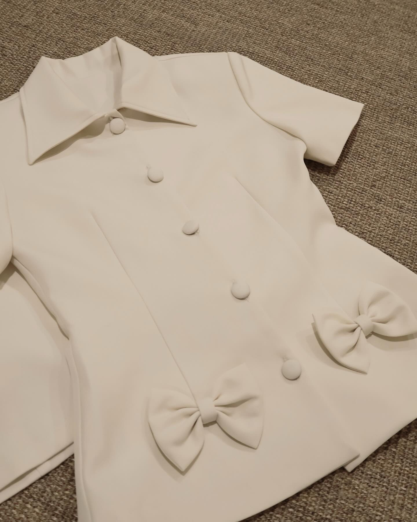 The.lookbook.select 的 Light Skirt Set 🖤新品優惠 2 件9折的特色是一件白色短袖上衣，有尖領、布料鈕扣，下擺附近有兩個裝飾蝴蝶結，平鋪在有紋理的表面上。.