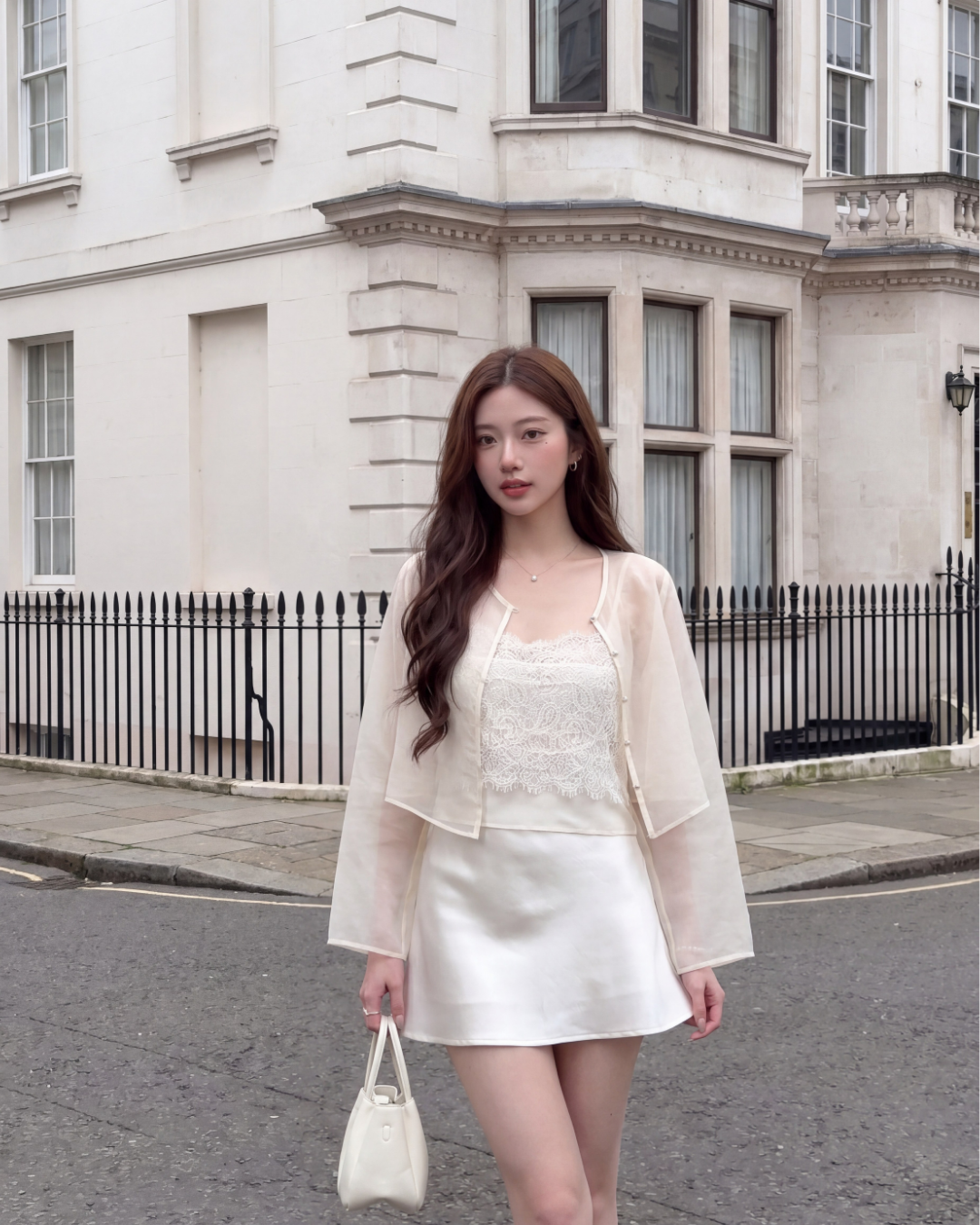 一位女士穿著 the.lookbook.select 的 Chiffon Arch Duo (Cami & Jacket) 純奶油色新品優惠2件9折，站在一棟有大窗戶的白色建築物旁，手上拿著一個小手包。.