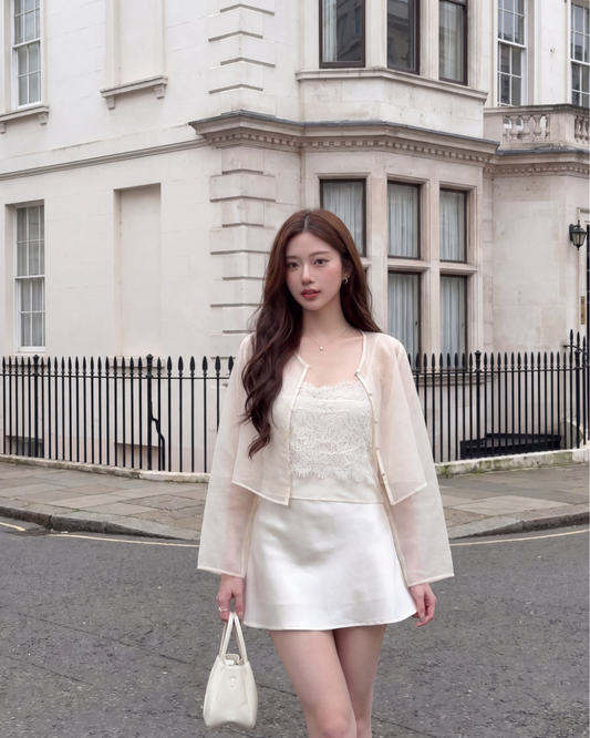一位女士穿著 the.lookbook.select 的 Chiffon Arch Duo (Cami & Jacket) 純奶油色新品優惠2件9折，站在一棟有大窗戶的白色建築物旁，手上拿著一個小手包。.