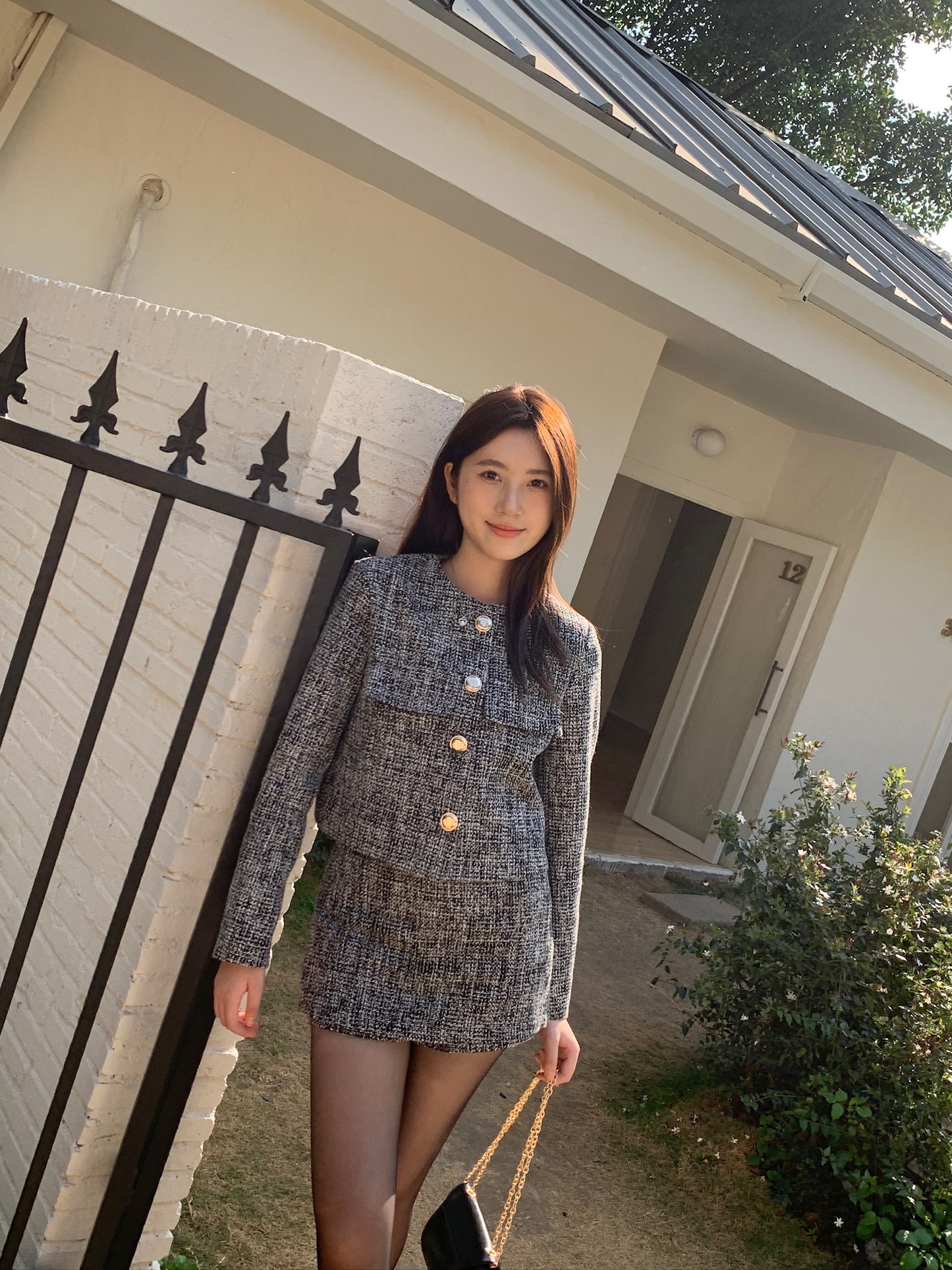 一個人站在一座白房子外面，穿著 the.lookbook.select 的 This Girl Tweed Set，手裡拿著一個帶鏈條帶的黑色錢包。.