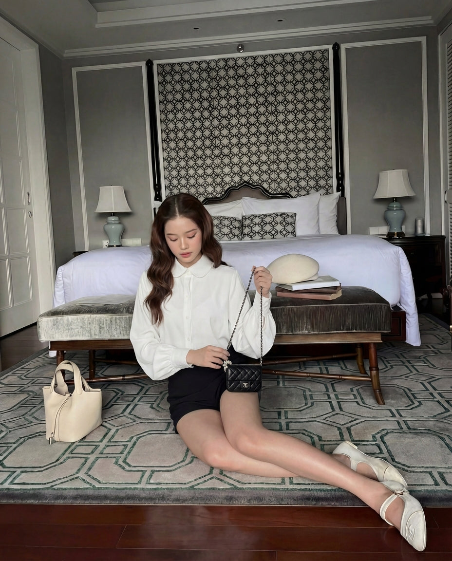 一位女士穿著 the.lookbook.select 的 Puff Sleeve Shirt 🖤新品優惠2件9折和黑色短褲，坐在臥室的地板上，背景是黑色的小皮包、床和兩張邊桌。.