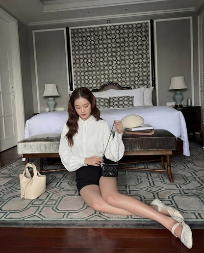 一位女士穿著 the.lookbook.select 的 Puff Sleeve Shirt 🖤新品優惠2件9折和黑色短褲，坐在臥室的地板上，背景是黑色的小皮包、床和兩張邊桌。.