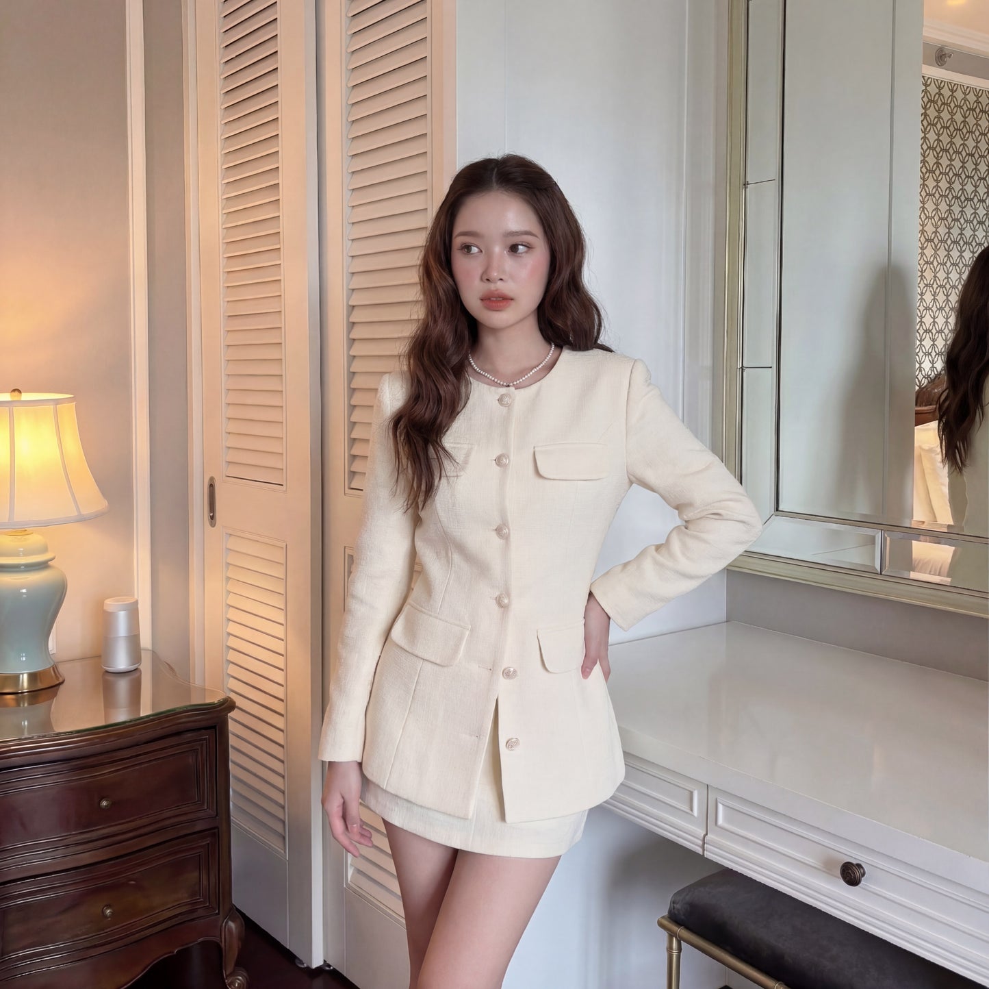一位女士站在室內，一手扶著臀部，身穿 the.lookbook.select 的 All White Set 🖤新品優惠 2 件 9 折 - 奶油色鈕扣外套和裙子，站在桌子和鏡子旁。.