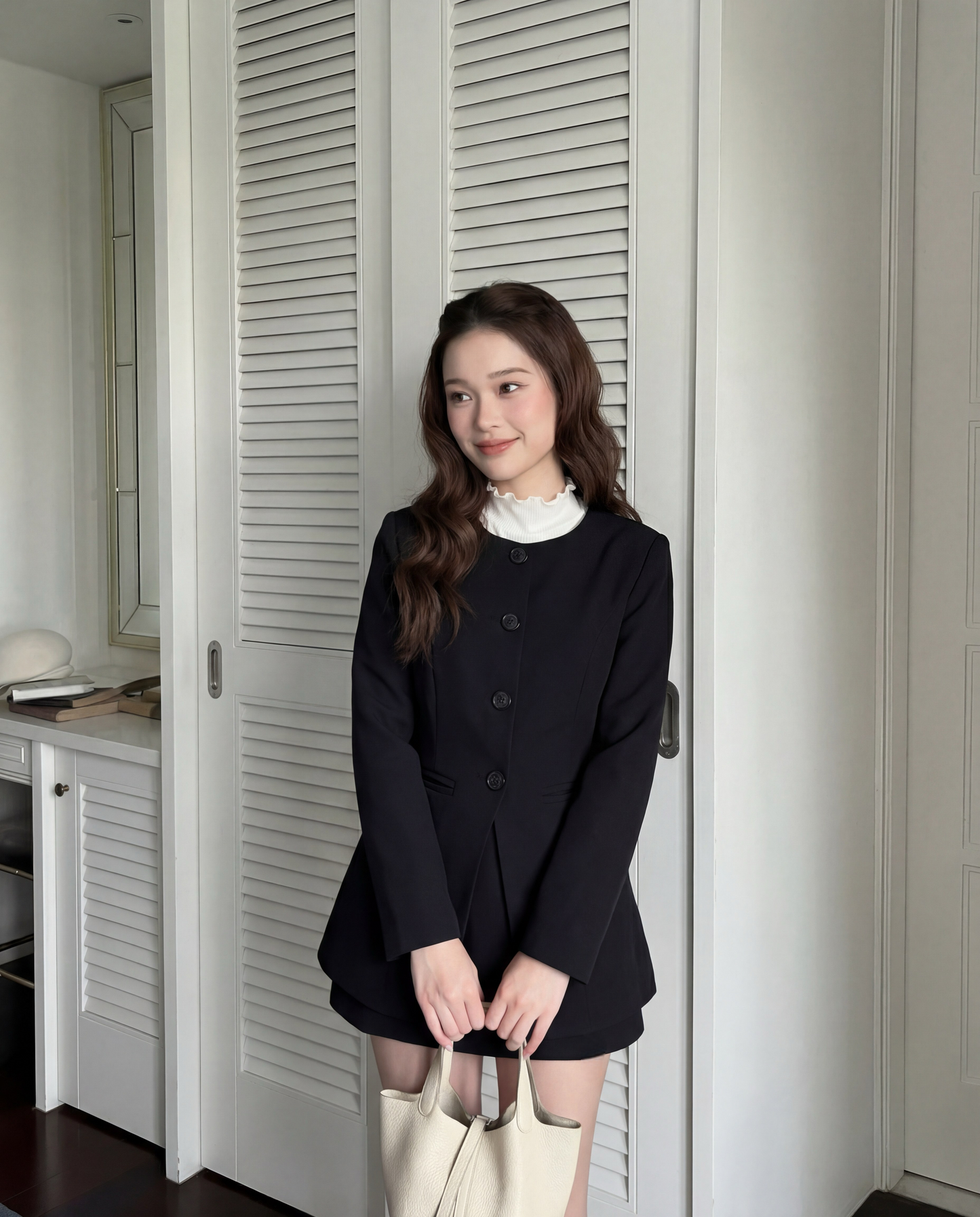 一位披著波浪長髮的女性，身穿the.lookbook.select的All Black Set (Coat & Pants)🖤新品優惠2件9折和白色上衣，手拿乳白色手提包，站在室內緊閉的白色衣櫃門前。.