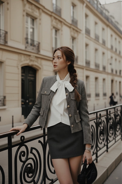 在米色建築林立的城市街道上，一位身穿 the.lookbook.select 的經典灰色粗花呢外套的女士站在裝飾性欄杆旁，她穿著白色蝴蝶結上衣和黑色短裙。.