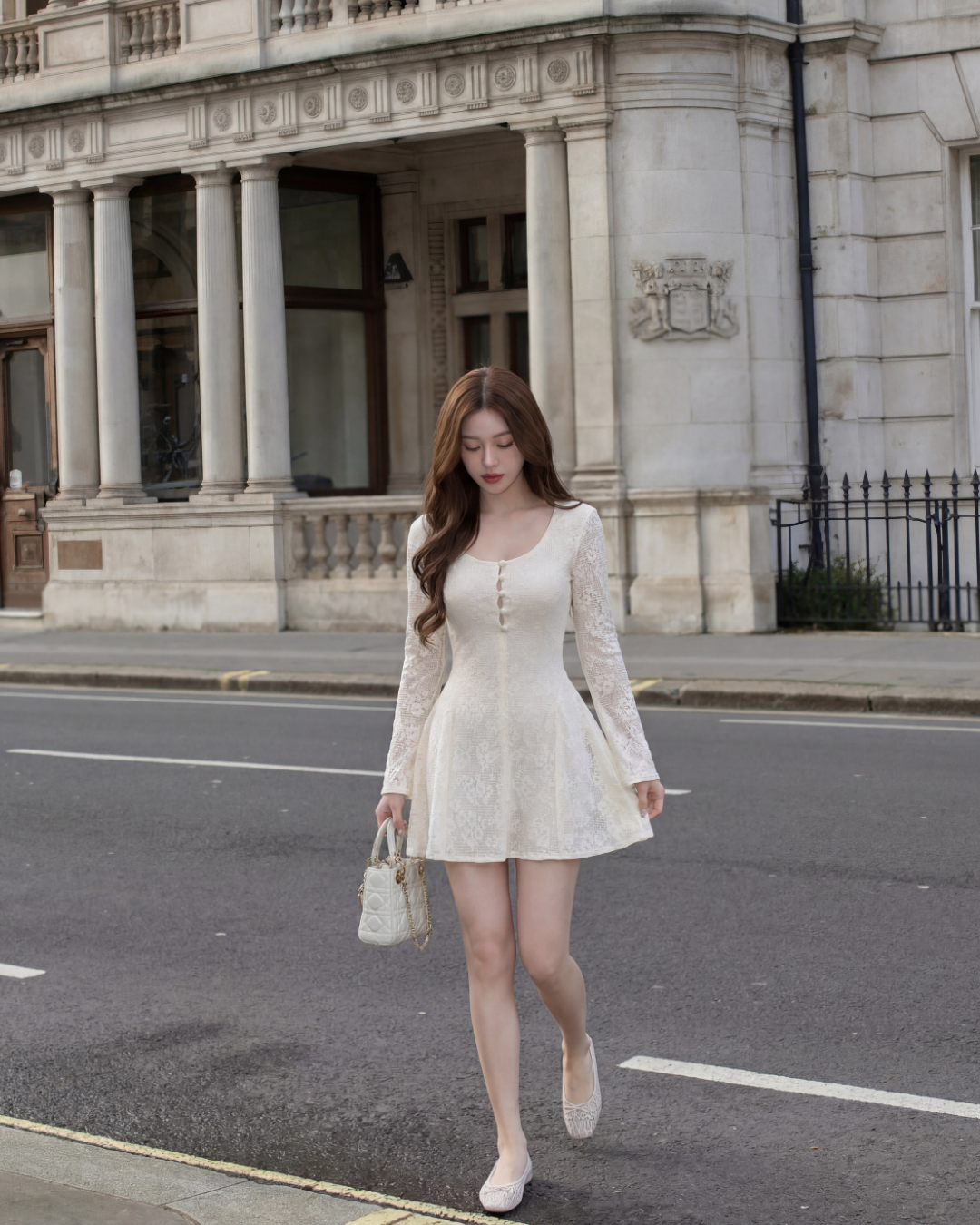 一位女士穿著 the.lookbook.select 的 Verse 蕾絲迷你裙，拎著小手包走在石造建築前的城市街道上。.