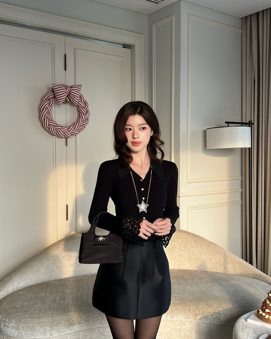 一位女士站在室內，身穿 the.lookbook.select 的蕾絲針織上衣❄️，搭配一件短裙，手拿黑色手提包。她身後那扇緊閉的雙門上掛著一個花圈。.