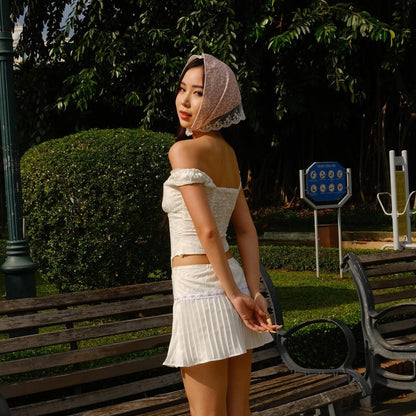 一名身穿 the.lookbook.select 的 Amour Set 的人站在公園裡的長凳和樹籬旁，其中包括一件飾有小花刺繡的白色露肩上衣和一條百褶裙。