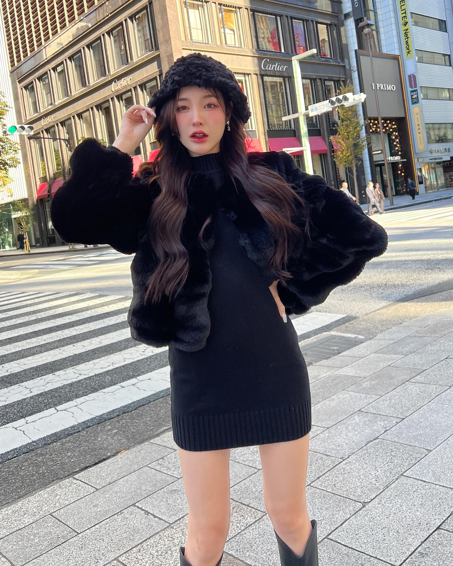 一位穿著 the.lookbook.select 的棉花糖皮草大衣 (附帽子) 的女性，自信地站在城市街道的人行道旁，身後是豪華商店的招牌。.