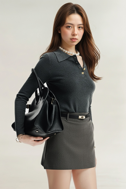 素色背景下，一位女士面朝前方，身穿 the.lookbook.select 的灰色 Luna 迷你裙、深色長袖上衣和珍珠項鍊，肩上挎著一個黑色大手提包。.