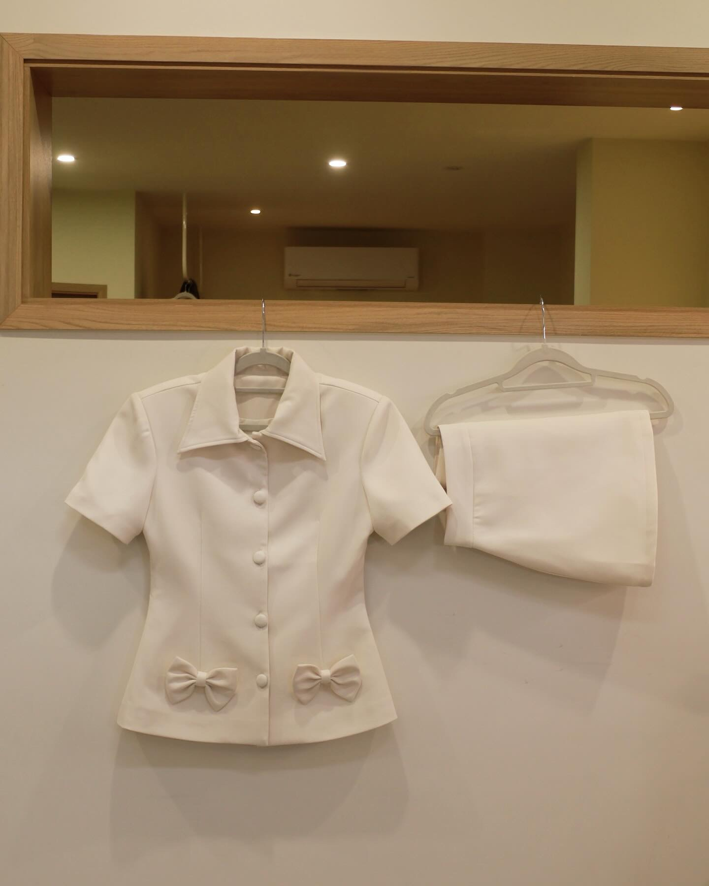 The.lookbook.select推出的Light Skirt Set，以白色短袖鈕扣上衣搭配蝴蝶結點綴的短裙為主，掛在木框鏡子下方的掛牆架上。.