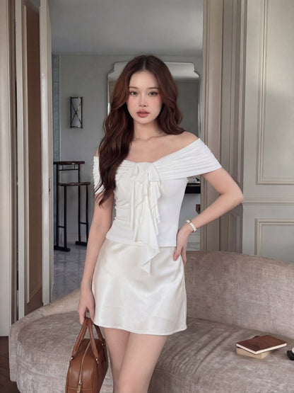 一位女士穿著 the.lookbook.select 的 Petal Bardot Top，拎著棕色手提包站在時尚的房間裡。她身旁的米色沙發上放著一本合上的筆記本。.