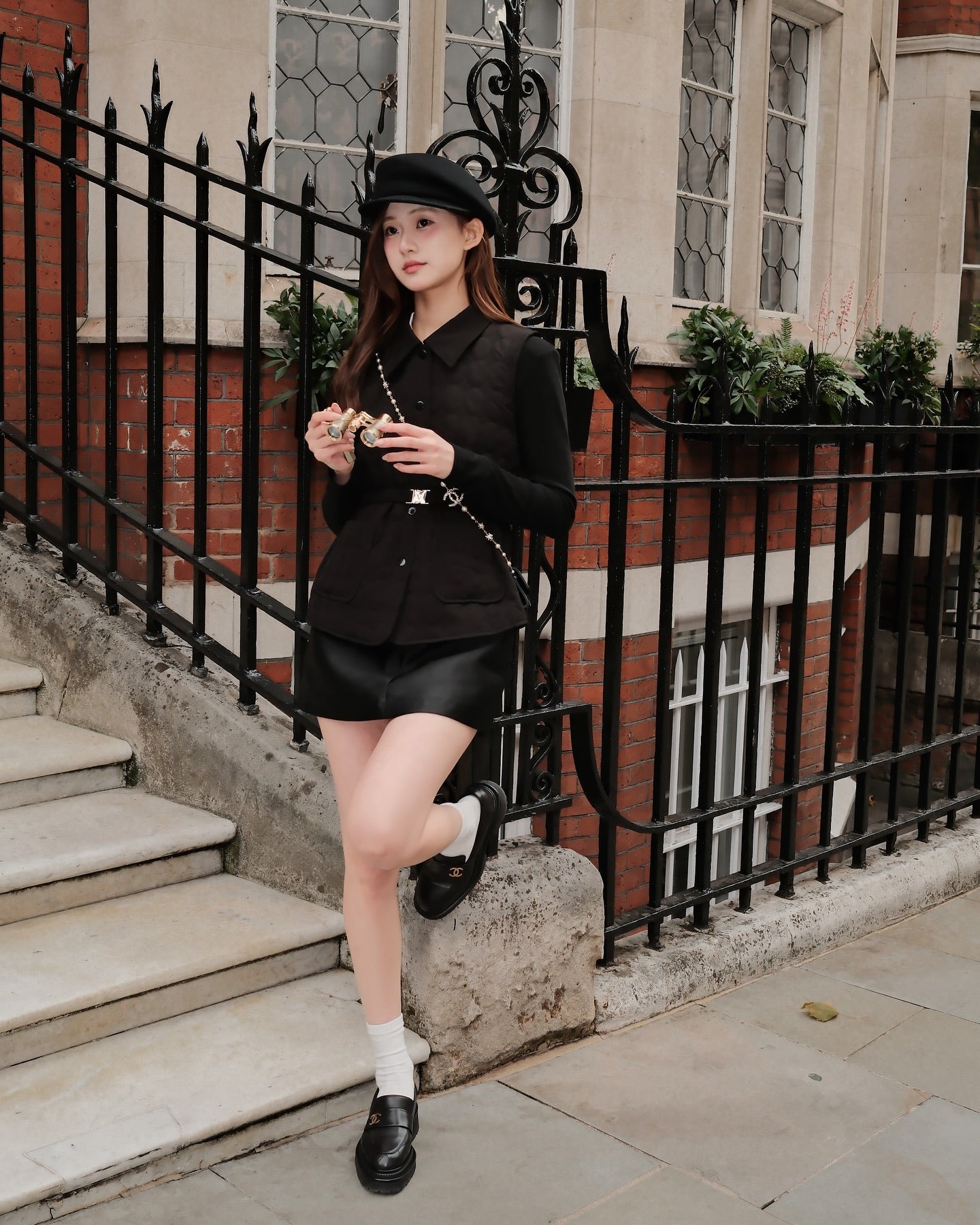 一個穿著黑色衣服，戴著帽子的女人，站在樓梯和鐵柵欄旁，拿著食物看著一旁，身穿the.lookbook.select 的Diana Vest Coat 🖤，展現法式精緻風格，輕盈保暖又優雅。.