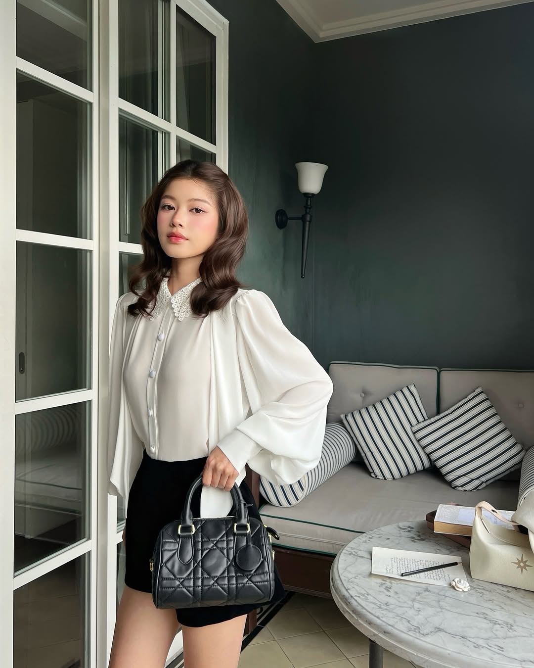 一位女士穿著 the.lookbook.select 的 Heart White Shirt ❄️ 新品優惠和黑色短褲，在室內拿著一個黑色手提包。她身後的沙發上有條紋抱枕，圓桌上則擺放著各式各樣的物品。.