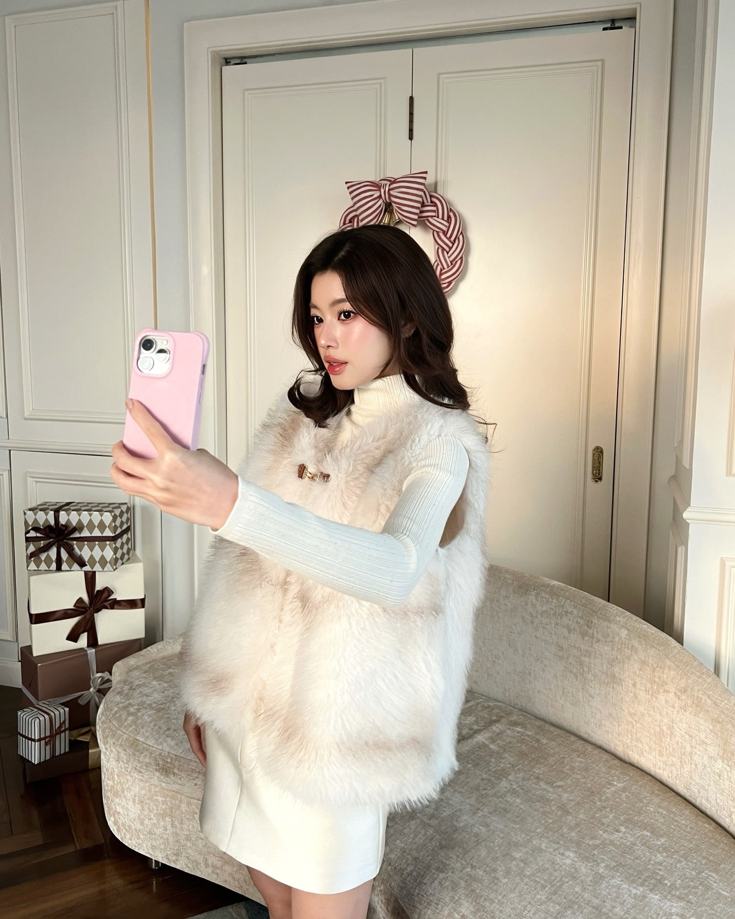 一位身穿.lookbook.select La Neige Fur Vest ❄️ 新品優惠、手持粉紅色手機的女士，在佈滿禮盒和中性裝潢的房間裡自拍，展現時尚又保暖的冬日穿搭。.