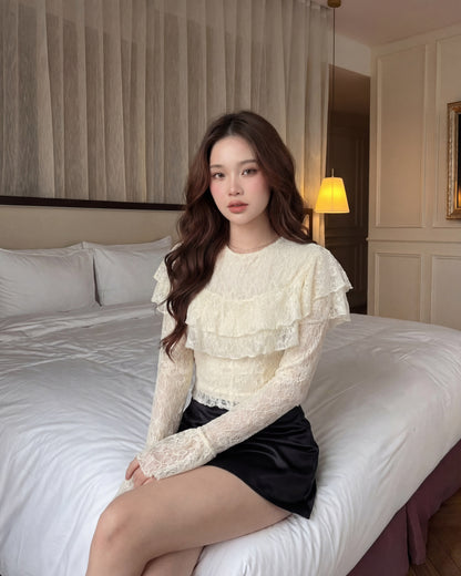 一位留著長捲髮的女子坐在燈光柔和的房間裡那張整理得整整齊齊的床上，身穿 the.lookbook.select 的「春季繆斯」蕾絲上衣與黑色裙子，散發出一股優雅而浪漫的氛圍。.