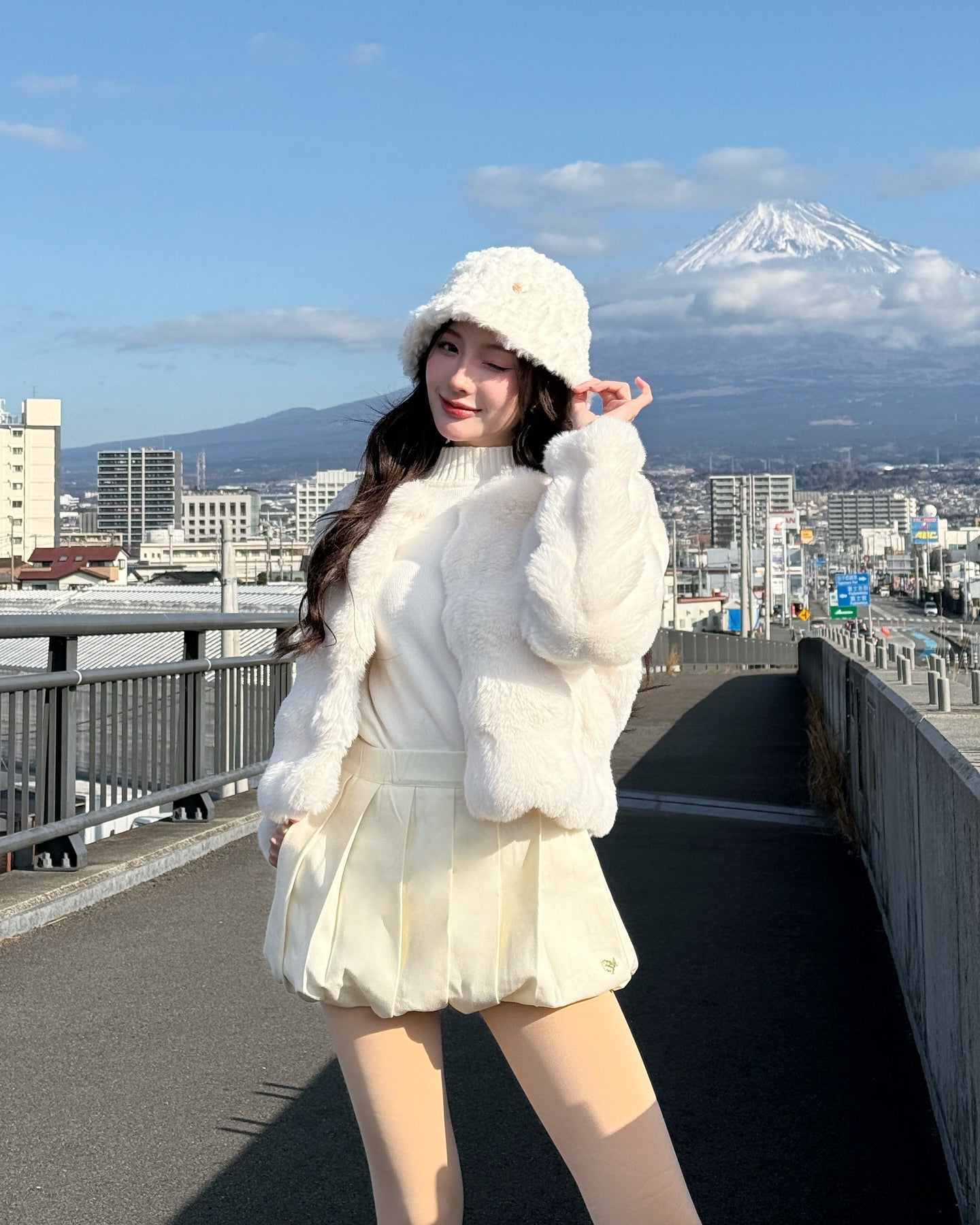 一位女士在城市人行道上為 the.lookbook.select 的棉花糖皮草大衣 (配帽子) 當模特，背景是多雲的天空下的富士山。.