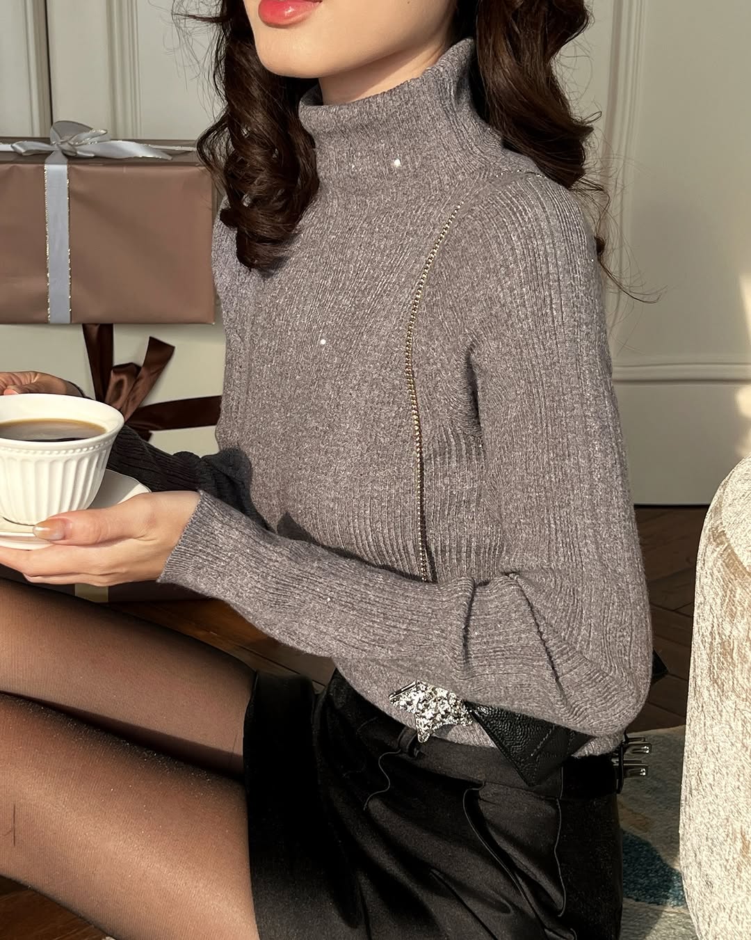 一位身穿 the.lookbook.select's Twinkle Knit Top❄️新品優惠和黑色短裙的女士手持一杯咖啡，背景可見包裝好的禮物。.