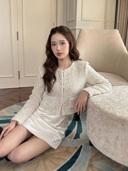 一名女子坐在一張米色椅子旁的地板上，身穿 the.lookbook.select 的「月光開襟衫」與一條緞面白色裙子，身處光線充足、優雅的房間中，展現法式風情。.