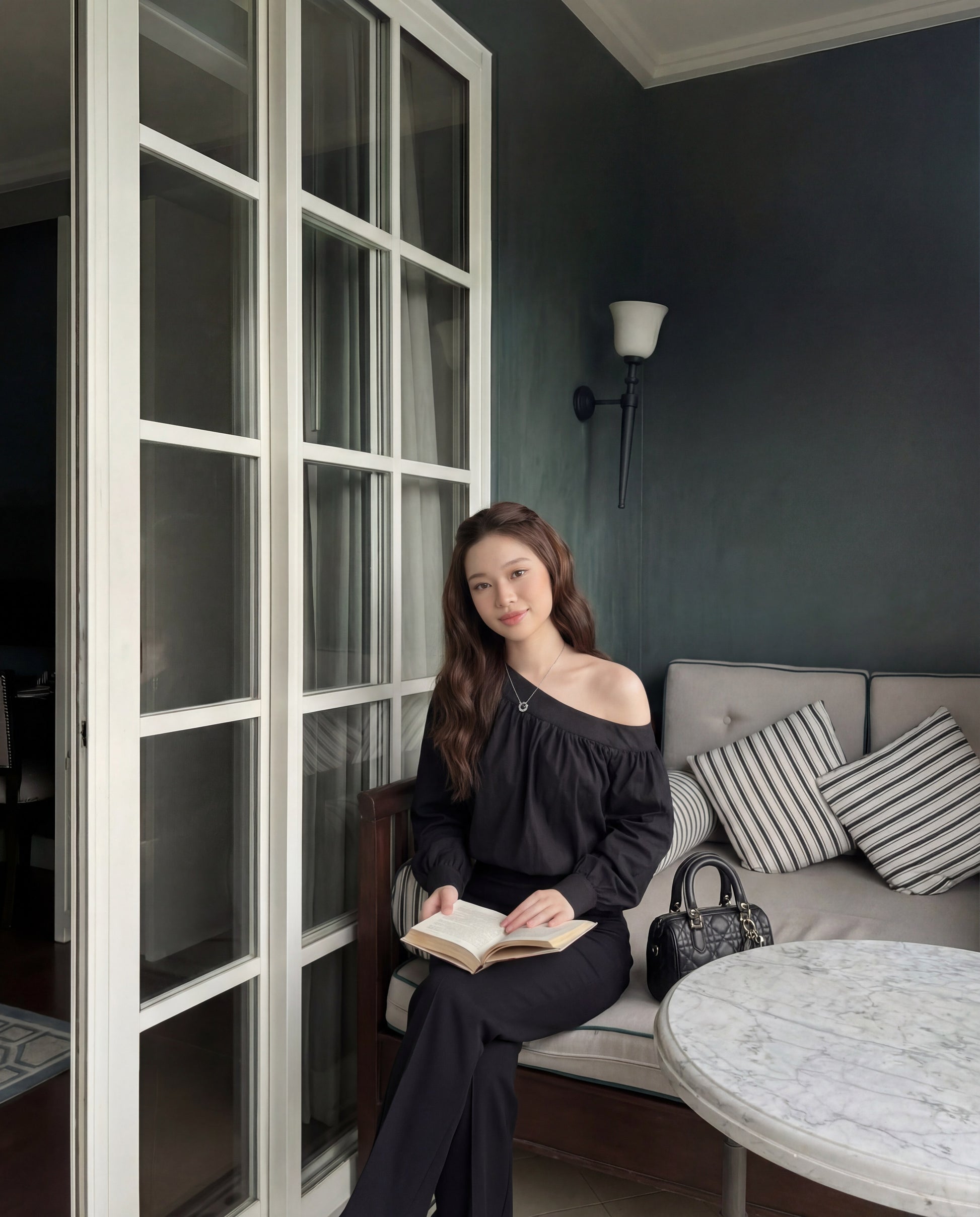 一位身穿 the.lookbook.select 黑色 Lou 垂褶衫的女士坐在大理石圓桌旁的軟墊長椅上，手持一本打開的書，身旁是條紋抱枕和黑色手提包。.
