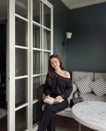 一位身穿 the.lookbook.select 黑色 Lou 垂褶衫的女士坐在大理石圓桌旁的軟墊長椅上，手持一本打開的書，身旁是條紋抱枕和黑色手提包。.