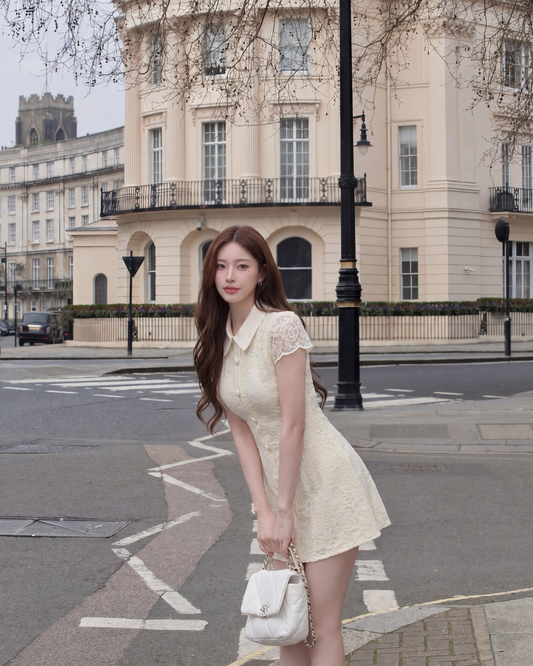 一位身穿 the.lookbook.select 惠靈頓蕾絲連衣裙的女士拿著一個白色手提包，站在城市街道上一棟有柱和陽台的大型米色建築前。.