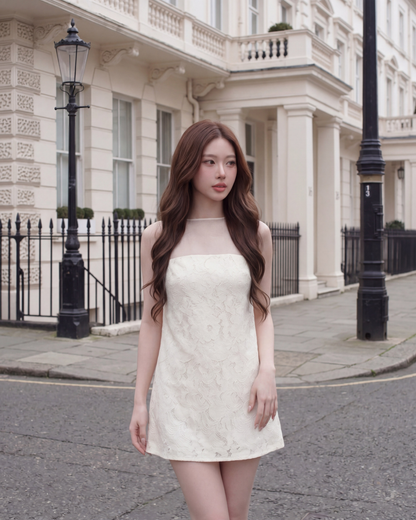 一位留著長棕髮的女子身穿由 the.lookbook.select 推出的「Elysee 蕾絲迷你洋裝」，佇立在一條兩旁盡是米色建築物的寧靜城市街道上。.