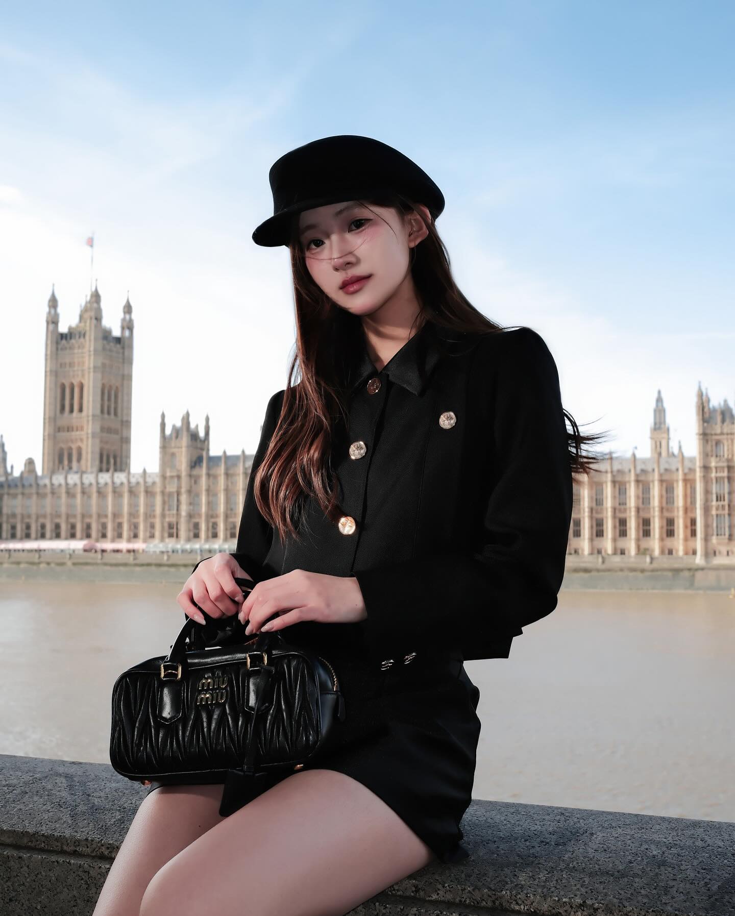 一位女士穿著 the.lookbook.select 的 Grace Little Jacket 🖤新品優惠至 12 月 5 日，戴著帽子，坐在石台上拿著黑色手提包，背景是威斯敏斯特宮和泰晤士河。.