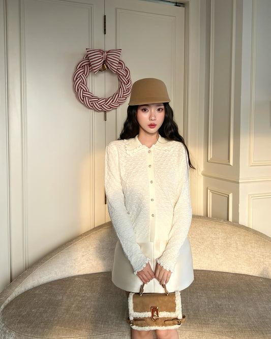 一位女士穿著 the.lookbook.select 的 Janelle 針織開襟衫❄️ 優惠新品和一頂米色帽子，拎著一個小手包站在弧形沙發旁；她身後的門上掛著一個花圈。.