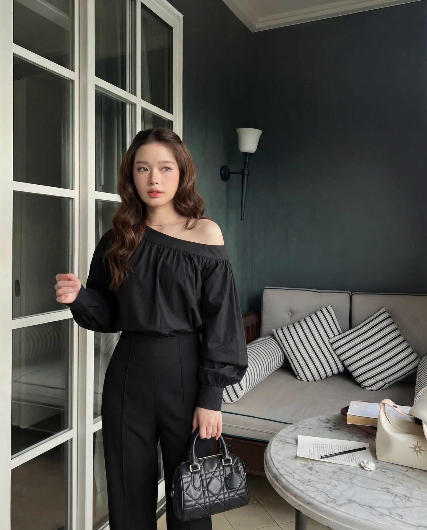 一位室內女士穿著 the.lookbook.select 的 Lou Drape 襯衫搭配黑色褲子，手拿黑色手提包。背景可見一張沙發、一張放有書本和錢包的桌子。.