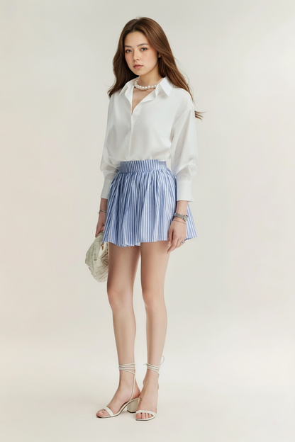 一位女士身穿 the.lookbook.select 的 Lady Oversized Shirt，搭配藍白條紋短裙、白色綁帶涼鞋，手拿小包包，站在素色背景中。.