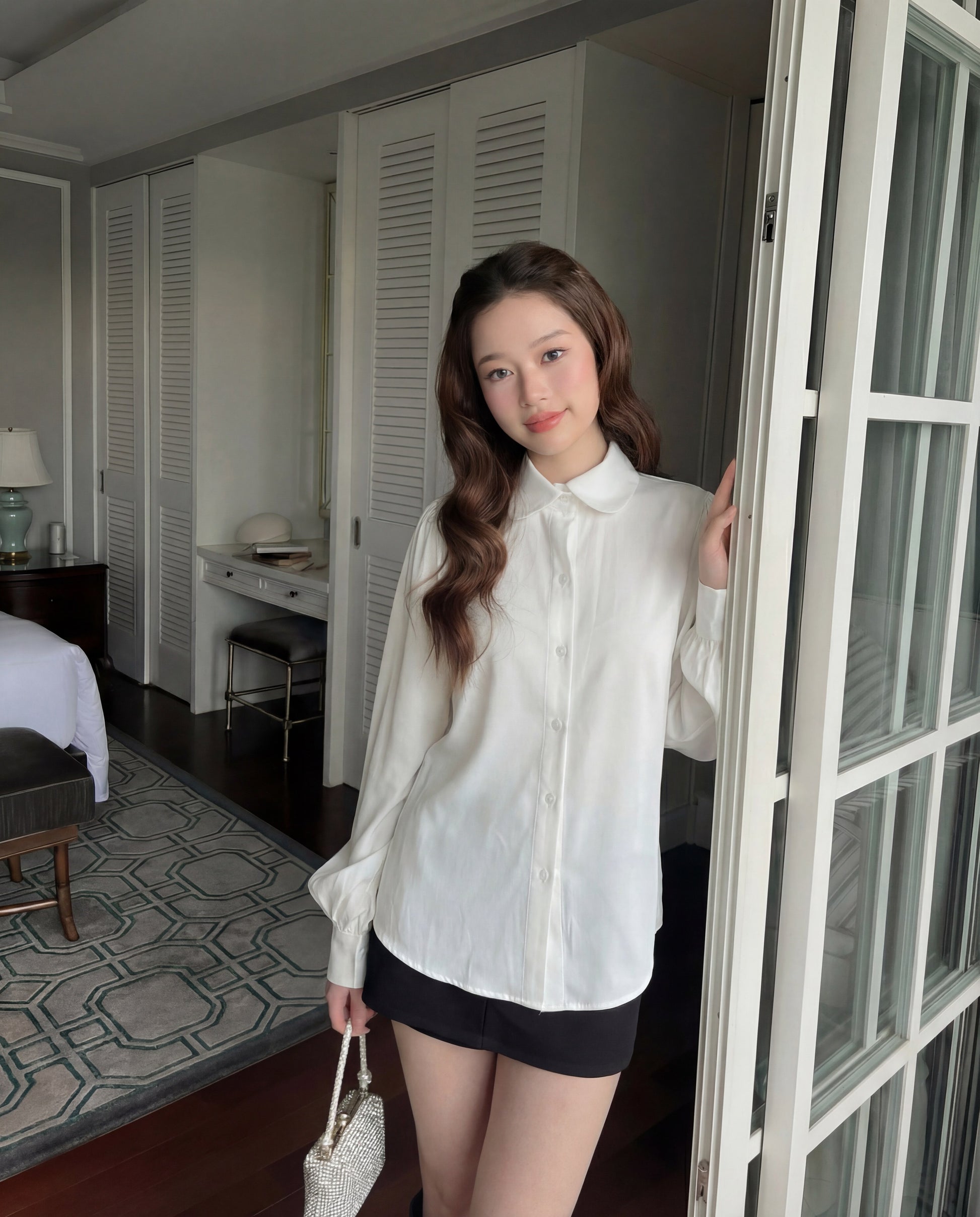 一位身穿 the.lookbook.select 白色 Puff Sleeve Shirt 和黑色短裙的女士站在房間裡，拿著手提包，靠在玻璃門上。.